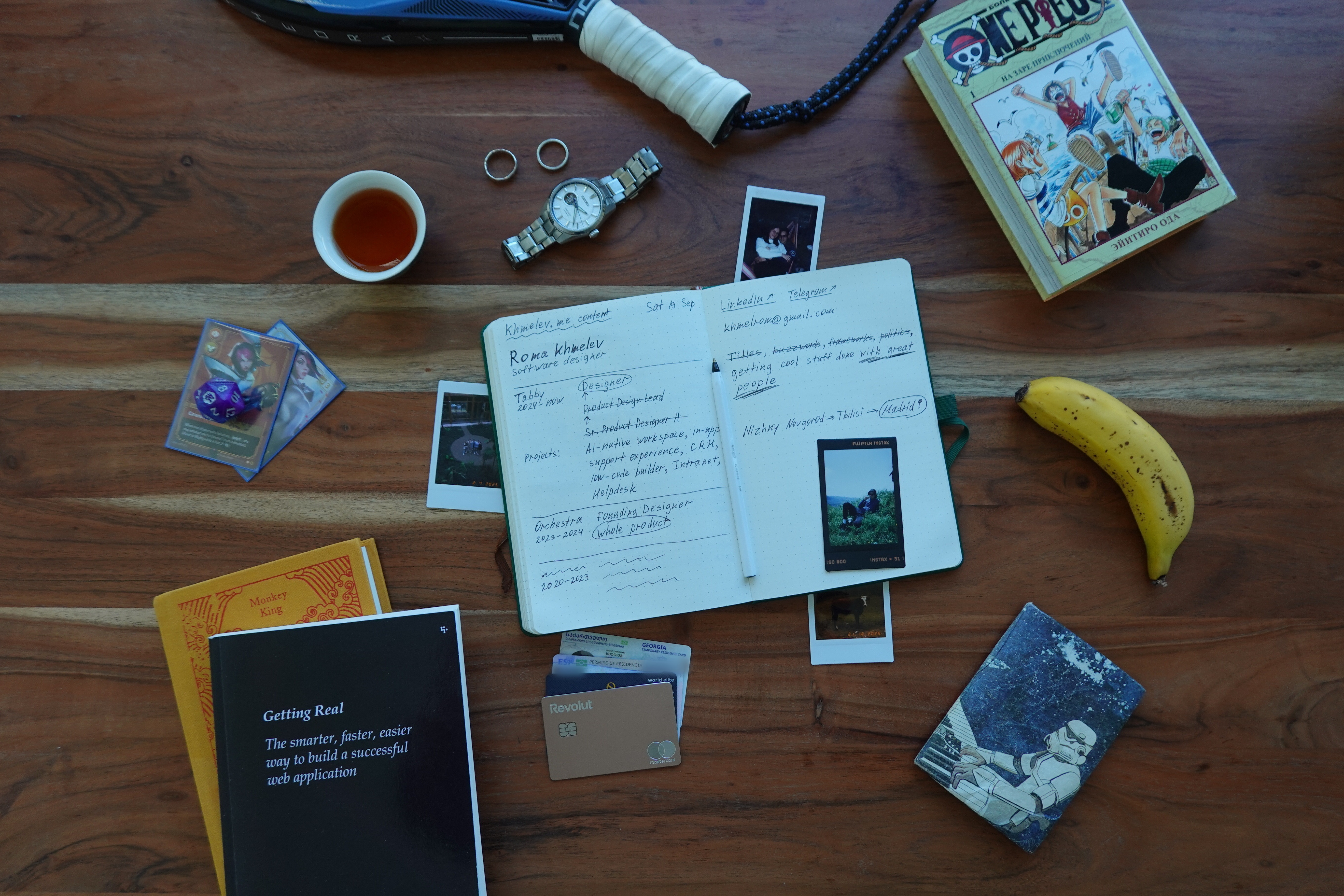 A desk that tells a lot about me: my notebook with the khmelev.me story — designer at Tabby, ex-Orchestra — surrounded by a padel racket, a Seiko watch, One Piece manga, Getting Real, tea, and a banana.