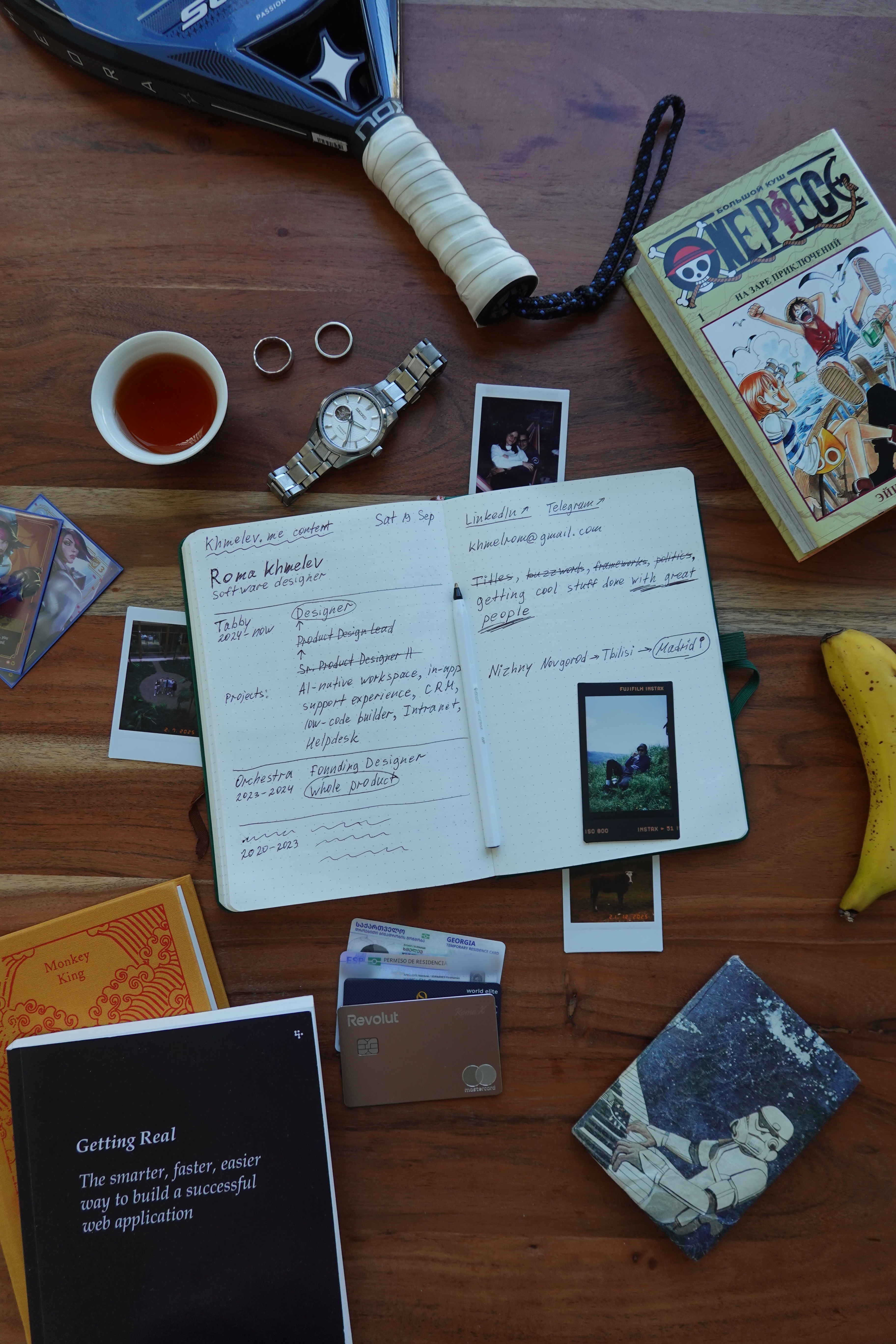 A desk that tells a lot about me: my notebook with the khmelev.me story — designer at Tabby, ex-Orchestra — surrounded by a padel racket, a Seiko watch, One Piece manga, Getting Real, tea, and a banana.
